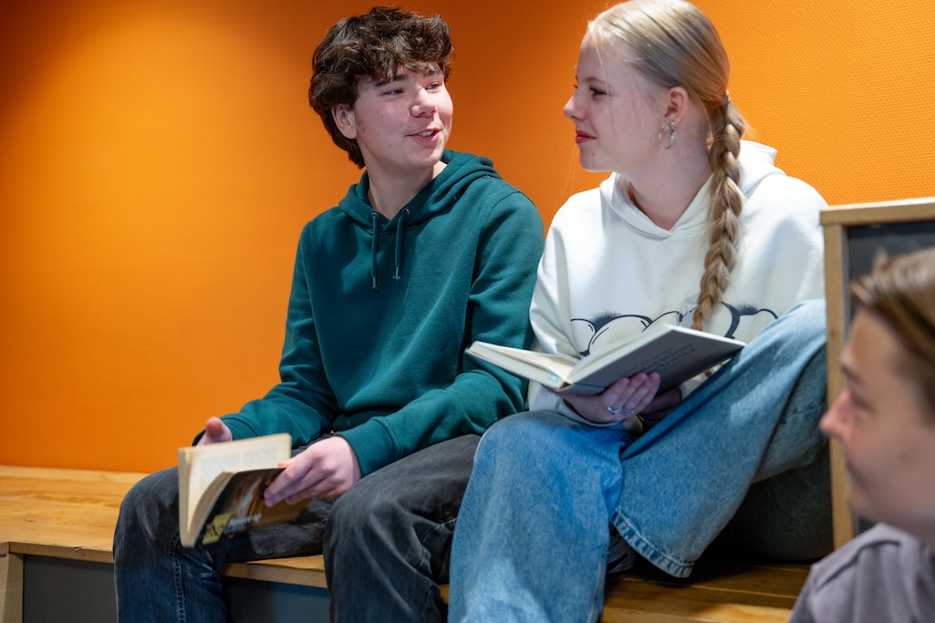 Twee leerlingen kijken elkaar aan en hebben elk een opengeslagen boek vast, zitten op een verhoging.